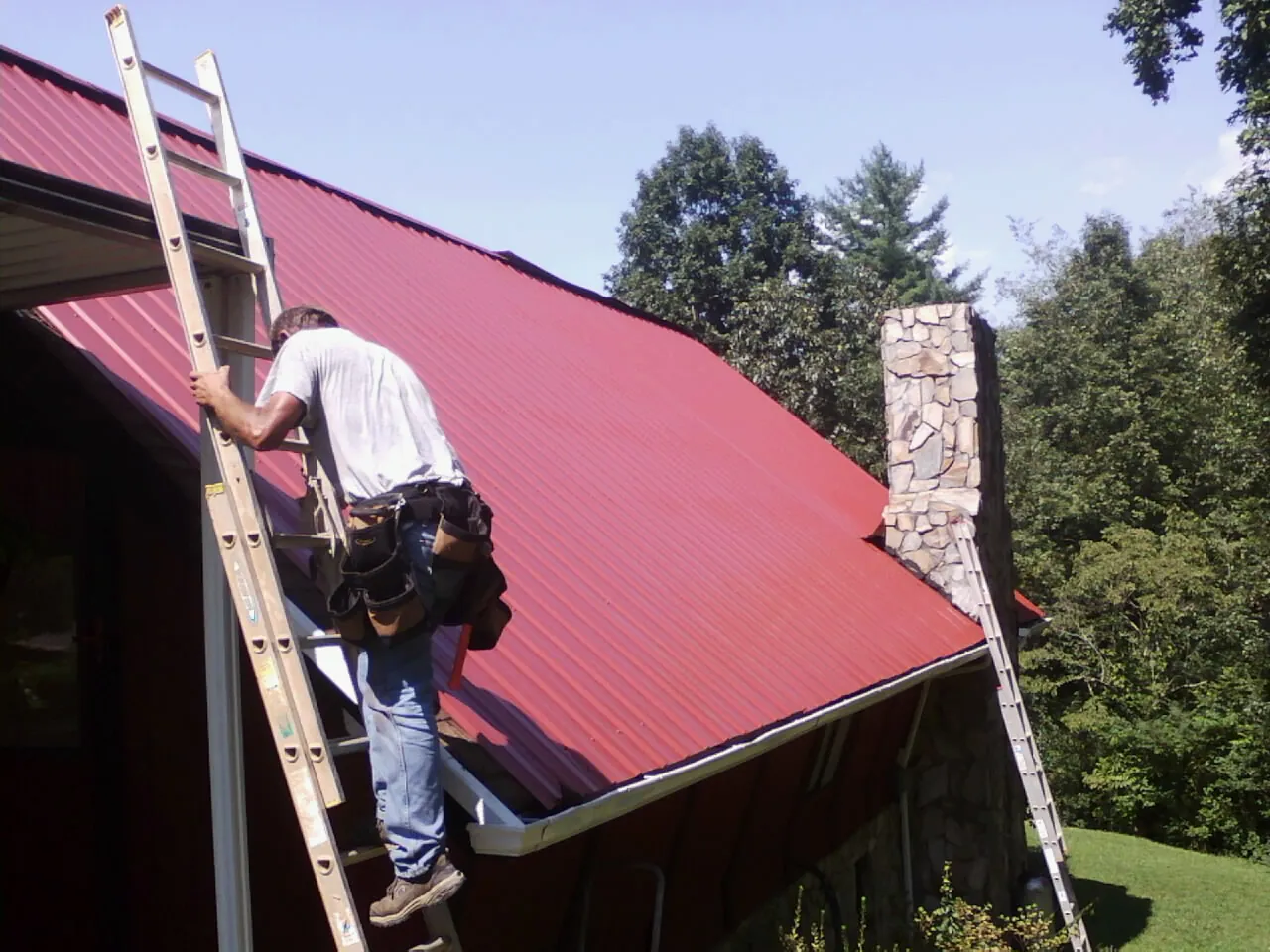Roofer on a ladder beside a red metal roof and stone chimney — Soffit & Fascia Repair by Roofing Brothers in Kelly